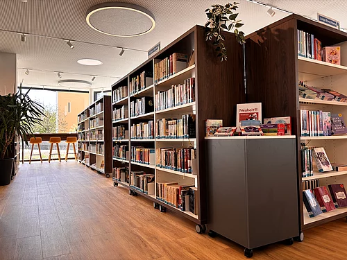 Mehrere hohe Bücherregale prägen den offenen Publikumsbereich der Stadtbücherei. Zwischen den Regalen stehen moderne Sitzmöglichkeiten und Pflanzen. Große Fenster lassen viel Tageslicht herein und schaffen eine freundliche, ruhige Lern- und Leseatmosphäre.