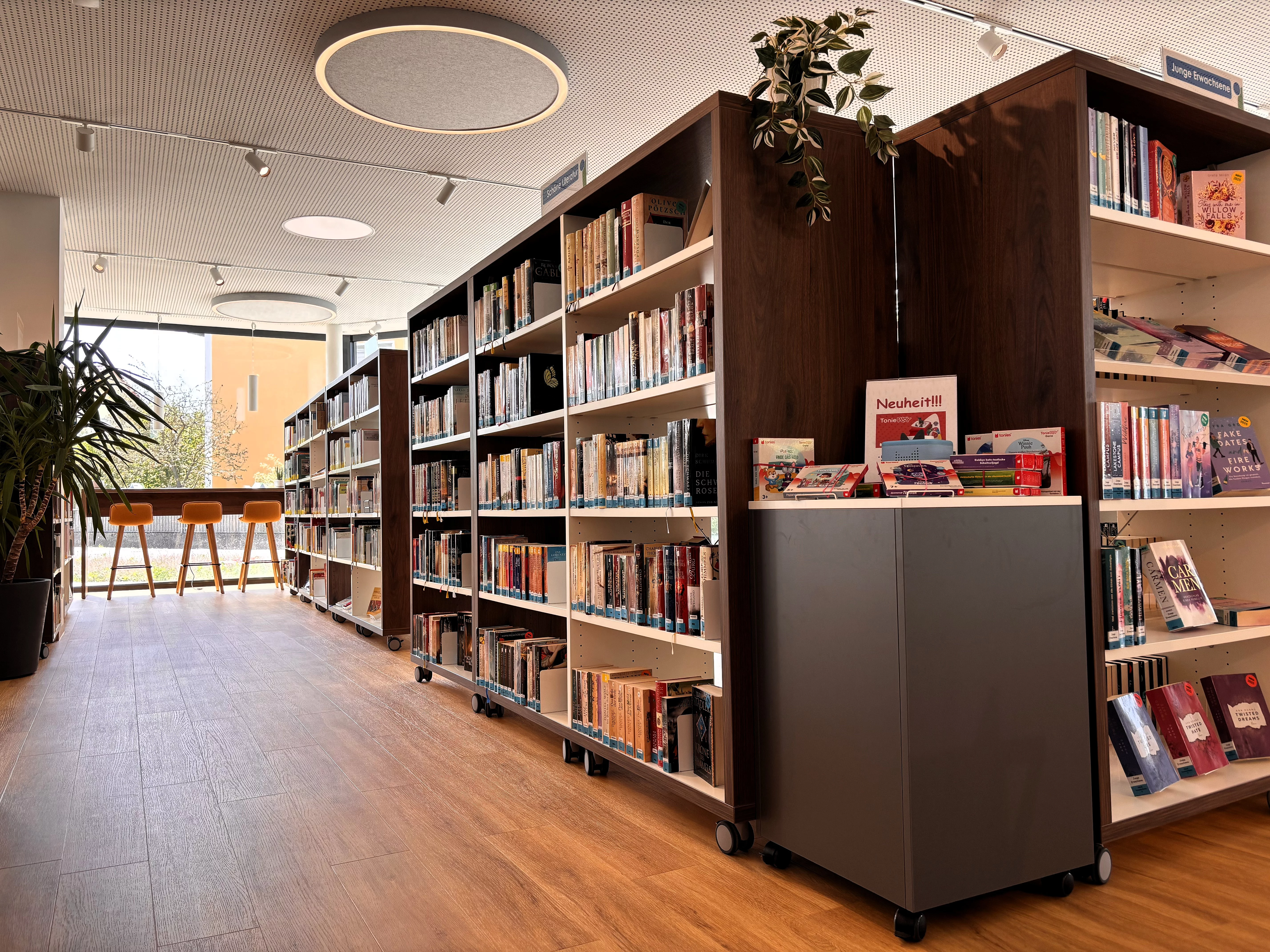 Mehrere hohe Bücherregale prägen den offenen Publikumsbereich der Stadtbücherei. Zwischen den Regalen stehen moderne Sitzmöglichkeiten und Pflanzen. Große Fenster lassen viel Tageslicht herein und schaffen eine freundliche, ruhige Lern- und Leseatmosphäre.