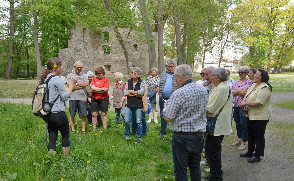 Das Foto zeigt eine Gruppe von Menschen, die im Freien an einer Führung oder einem Vortrag teilnimmt. Eine Frau mit Rucksack spricht vor der Gruppe und hält etwas in der Hand, vermutlich eine Pflanze. Die Teilnehmenden hören interessiert zu. Im Hintergrund sind Bäume und die Ruine eines alten Gebäudes zu sehen, was auf einen Park oder eine historische Anlage hinweist.