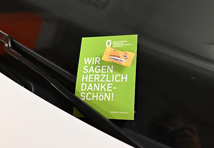 Eine grüne Dankeskarte des Zweckverbands Kommunale Verkehrssicherheit Oberpfalz steckt hinter dem Scheibenwischer eines Autos und trägt den Schriftzug ‚Wir sagen herzlich Dankeschön!‘ sowie den Hinweis auf eine kleine Aufmerksamkeit für korrekt parkende Verkehrsteilnehmer.