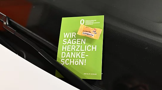 Eine grüne Dankeskarte des Zweckverbands Kommunale Verkehrssicherheit Oberpfalz steckt hinter dem Scheibenwischer eines Autos und trägt den Schriftzug ‚Wir sagen herzlich Dankeschön!‘ sowie den Hinweis auf eine kleine Aufmerksamkeit für korrekt parkende Verkehrsteilnehmer.