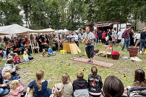 Ein Gaukler tritt auf einer kleinen Freifläche vor zahlreichen Kindern und Erwachsenen auf. Die Zuschauer sitzen oder stehen im Halbkreis und verfolgen aufmerksam die Vorführung. Im Hintergrund befinden sich Marktstände und Zelte des Mittelalterfestes.