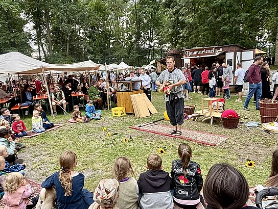 Ein Gaukler tritt auf einer kleinen Freifläche vor zahlreichen Kindern und Erwachsenen auf. Die Zuschauer sitzen oder stehen im Halbkreis und verfolgen aufmerksam die Vorführung. Im Hintergrund befinden sich Marktstände und Zelte des Mittelalterfestes.