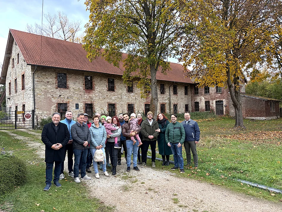 Gruppenfoto vom zweiten Neubürgerempfang der Stadt Teublitz im Herbst 2026. Eine Gruppe von Erwachsenen und zwei kleinen Kindern steht auf einem Kiesweg vor dem historischen Industriedenkmal „d’Schleif“ in Münchshofen. Die Teilnehmenden tragen herbstliche Jacken und Mäntel, zwei Erwachsene halten jeweils ein Kind auf dem Arm. Im Hintergrund ist das langgezogene historische Gebäude mit Natursteinfassade, zahlreichen Fenstern und rotem Ziegeldach zu sehen. Große Bäume mit gelblich-grünem Herbstlaub rahmen die Gruppe ein. Das Foto entstand bei Tageslicht während der gemeinsamen Besichtigung.