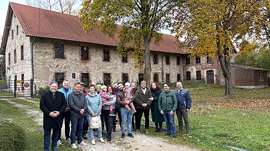 Gruppenfoto vom zweiten Neubürgerempfang der Stadt Teublitz im Herbst 2026. Eine Gruppe von Erwachsenen und zwei kleinen Kindern steht auf einem Kiesweg vor dem historischen Industriedenkmal „d’Schleif“ in Münchshofen. Die Teilnehmenden tragen herbstliche Jacken und Mäntel, zwei Erwachsene halten jeweils ein Kind auf dem Arm. Im Hintergrund ist das langgezogene historische Gebäude mit Natursteinfassade, zahlreichen Fenstern und rotem Ziegeldach zu sehen. Große Bäume mit gelblich-grünem Herbstlaub rahmen die Gruppe ein. Das Foto entstand bei Tageslicht während der gemeinsamen Besichtigung.