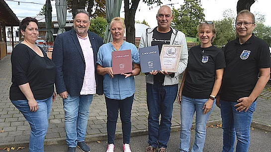 Sechs Personen stehen bei einer Ehrung vor einem Gasthaus und präsentieren zwei Urkunden, während die anderen Beteiligten gemeinsam für ein Gruppenfoto in die Kamera blicken.