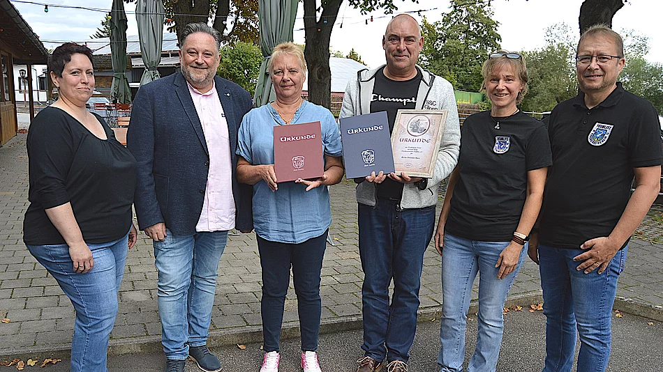 Sechs Personen stehen bei einer Ehrung vor einem Gasthaus und präsentieren zwei Urkunden, während die anderen Beteiligten gemeinsam für ein Gruppenfoto in die Kamera blicken.