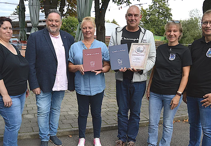 Sechs Personen stehen bei einer Ehrung vor einem Gasthaus und präsentieren zwei Urkunden, während die anderen Beteiligten gemeinsam für ein Gruppenfoto in die Kamera blicken.