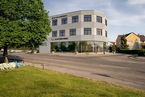 Ein modernes, zweigeschossiges Gebäude mit großen Fenstern und der Aufschrift „Stadtbücherei“ steht an einer Straße, umgeben von einem Baum, einer Grünfläche und benachbarten Wohnhäusern.