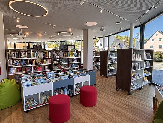 Heller, moderner Kinderbereich mit niedrigen Regalen, Sitzhockern und großen Fenstern.