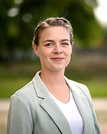 Eine Frau mit zusammengebundenem Haar und hellem Blazer blickt ruhig in die Kamera.
