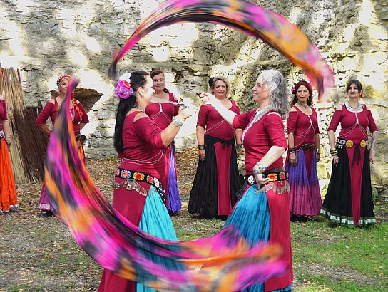 Mehrere Tänzerinnen in farbenfrohen mittelalterlich inspirierten Gewändern treten vor einer historischen Burgruine auf. Zwei Frauen schwingen lange bunte Schleier, während die übrigen Tänzerinnen die Vorführung begleiten. Einige Zuschauer verfolgen den Tanz vom Rand aus.