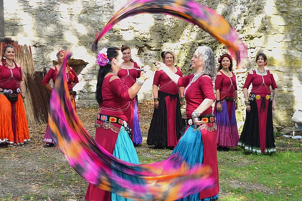 Mehrere Tänzerinnen in farbenfrohen mittelalterlich inspirierten Gewändern treten vor einer historischen Burgruine auf. Zwei Frauen schwingen lange bunte Schleier, während die übrigen Tänzerinnen die Vorführung begleiten. Einige Zuschauer verfolgen den Tanz vom Rand aus.