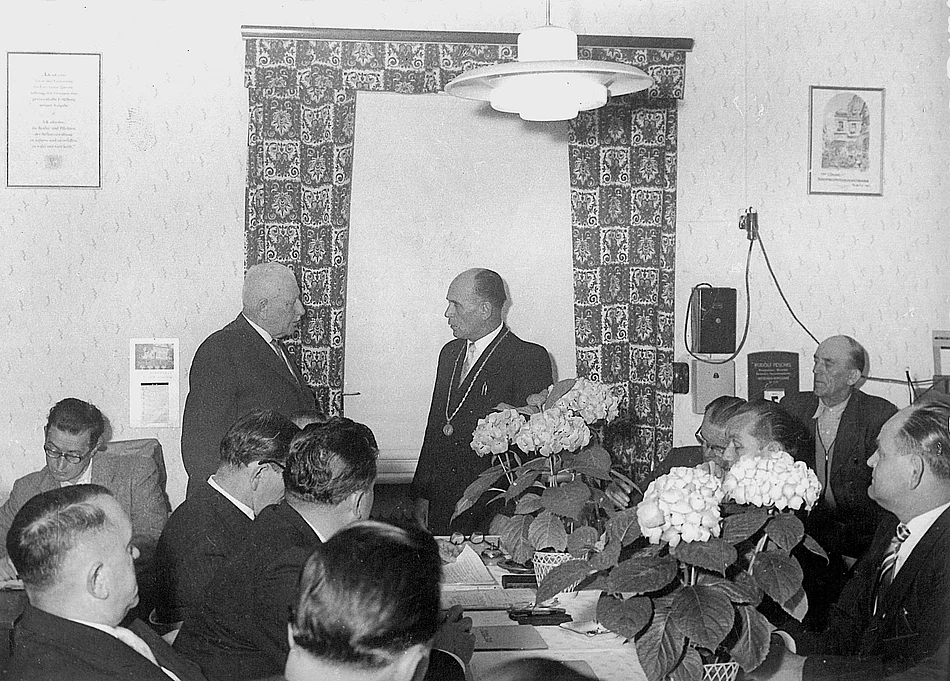 Auf dem Schwarzweißfoto ist eine Sitzung mit mehreren Männern in Anzügen an einem Tisch zu sehen, während zwei Männer im Mittelpunkt des Raumes miteinander sprechen – im Rahmen einer konstituierenden Stadtratssitzung im Jahr 1960.