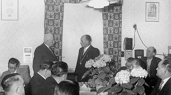 Auf dem Schwarzweißfoto ist eine Sitzung mit mehreren Männern in Anzügen an einem Tisch zu sehen, während zwei Männer im Mittelpunkt des Raumes miteinander sprechen – im Rahmen einer konstituierenden Stadtratssitzung im Jahr 1960.