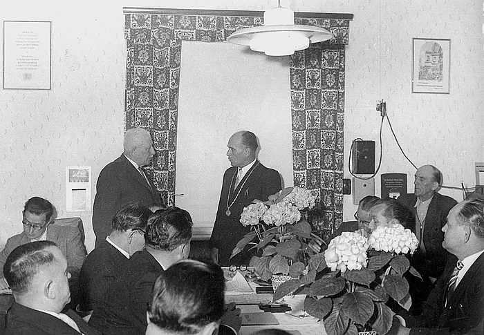 Auf dem Schwarzweißfoto ist eine Sitzung mit mehreren Männern in Anzügen an einem Tisch zu sehen, während zwei Männer im Mittelpunkt des Raumes miteinander sprechen – im Rahmen einer konstituierenden Stadtratssitzung im Jahr 1960.