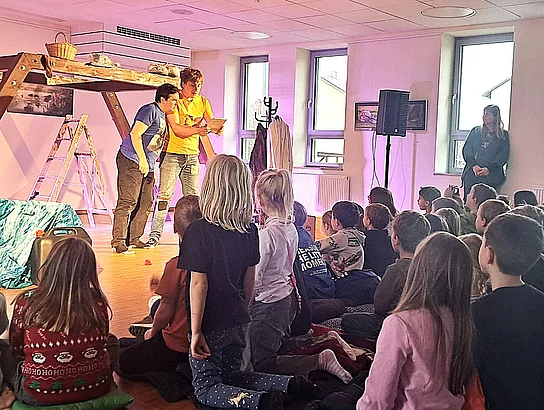 Viele Kinder sitzen gespannt auf dem Boden eines bunt beleuchteten Raumes und schauen zu, wie zwei Performer auf einer kleinen Bühne ein Stück aufführen, während eine erwachsene Person am Rand zusieht.