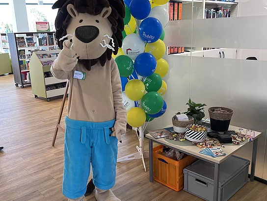 Ein Löwen-Maskottchen steht neben bunten Luftballons und einem Tisch mit kleinen Geschenken.