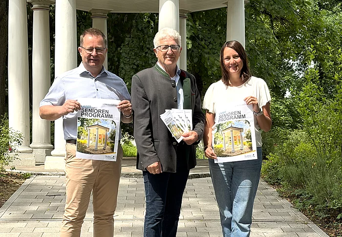 Drei Personen stehen auf einem gepflasterten Weg vor einem weißen Pavillon in einer grünen Parkanlage und präsentieren Broschüren des neuen Seniorenprogramms.