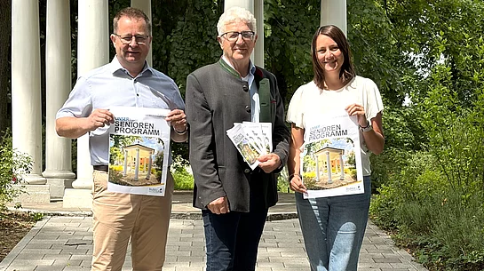 Drei Personen stehen auf einem gepflasterten Weg vor einem weißen Pavillon in einer grünen Parkanlage und präsentieren Broschüren des neuen Seniorenprogramms.