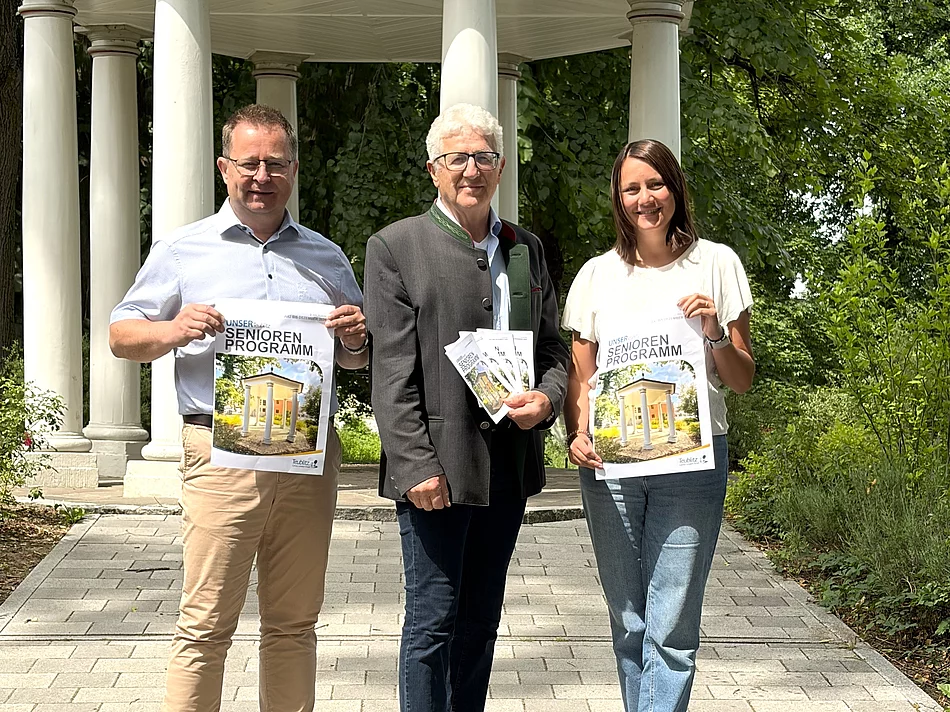 Drei Personen stehen auf einem gepflasterten Weg vor einem weißen Pavillon in einer grünen Parkanlage und präsentieren Broschüren des neuen Seniorenprogramms.