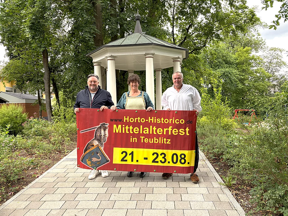 Drei Personen stehen auf einem gepflasterten Weg vor einem historischen Pavillon im Stadtpark und halten gemeinsam ein großes Werbebanner für das Mittelalterfest „Horto Historico“ in Teublitz. Das Banner zeigt das Veranstaltungslogo sowie den Termin vom 21. bis 23. August. Die beiden Personen rechts tragen mittelalterlich inspirierte Kleidung, während die Person links modern gekleidet ist. Umgeben ist die Szene von altem Baumbestand und viel Grün, wodurch die historische Kulisse besonders zur Geltung kommt.
