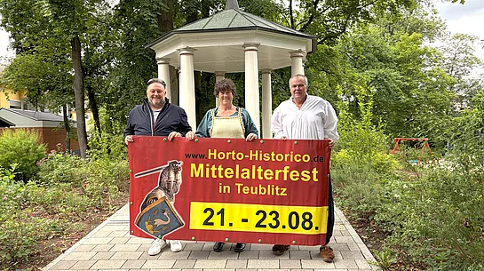 Drei Personen stehen auf einem gepflasterten Weg vor einem historischen Pavillon im Stadtpark und halten gemeinsam ein großes Werbebanner für das Mittelalterfest „Horto Historico“ in Teublitz. Das Banner zeigt das Veranstaltungslogo sowie den Termin vom 21. bis 23. August. Die beiden Personen rechts tragen mittelalterlich inspirierte Kleidung, während die Person links modern gekleidet ist. Umgeben ist die Szene von altem Baumbestand und viel Grün, wodurch die historische Kulisse besonders zur Geltung kommt.