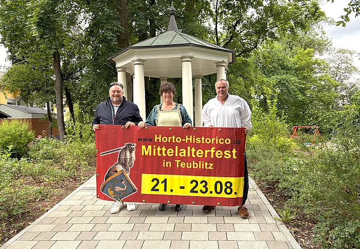 Drei Personen stehen auf einem gepflasterten Weg vor einem historischen Pavillon im Stadtpark und halten gemeinsam ein großes Werbebanner für das Mittelalterfest „Horto Historico“ in Teublitz. Das Banner zeigt das Veranstaltungslogo sowie den Termin vom 21. bis 23. August. Die beiden Personen rechts tragen mittelalterlich inspirierte Kleidung, während die Person links modern gekleidet ist. Umgeben ist die Szene von altem Baumbestand und viel Grün, wodurch die historische Kulisse besonders zur Geltung kommt.