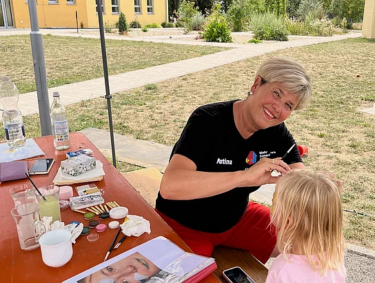 Eine Frau bemalt das Gesicht eines kleinen Kindes an einem Bastel- und Schminktisch im Außenbereich des Mehrgenerationenhauses.