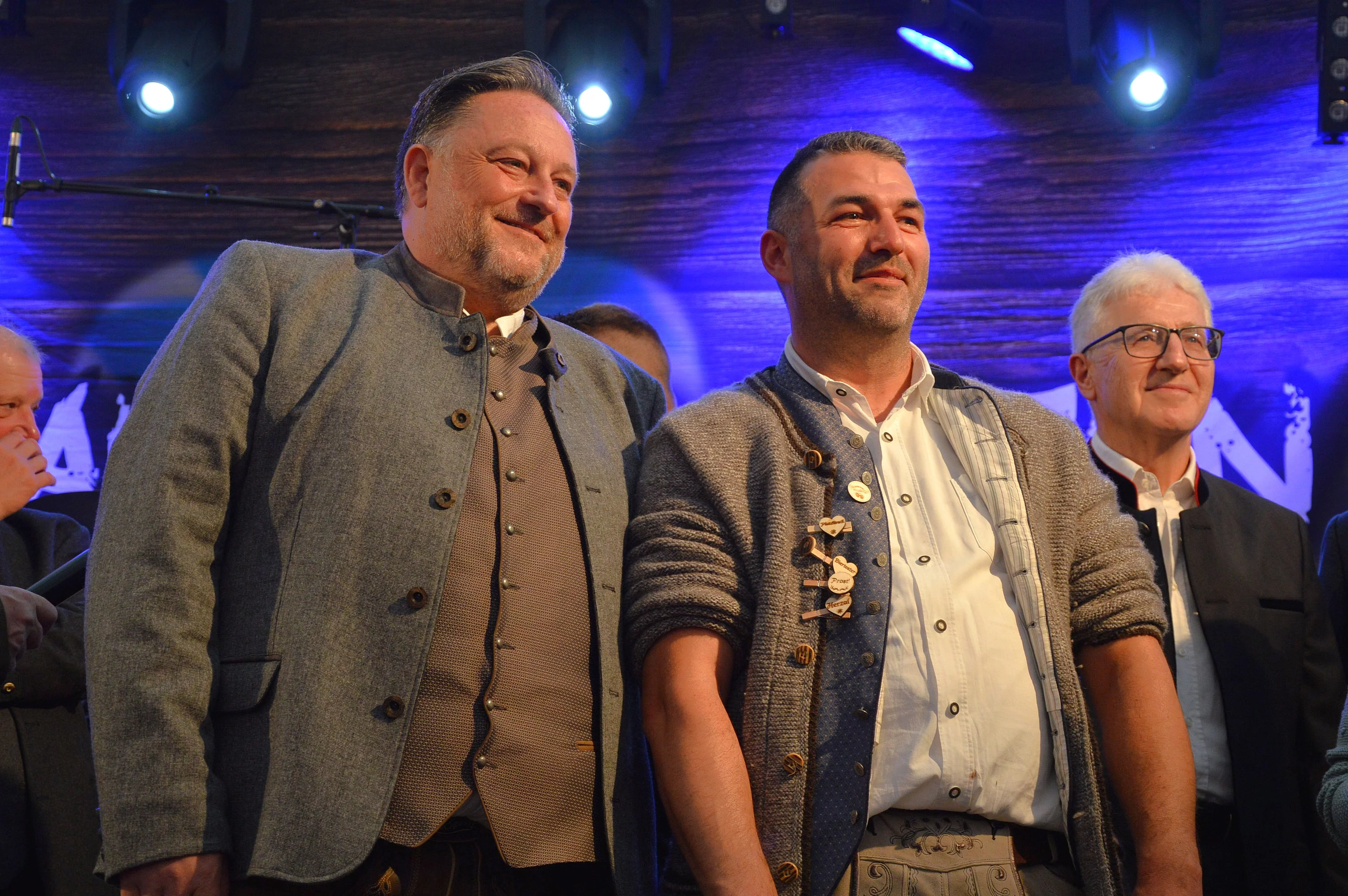 Drei Männer in Trachtenkleidung stehen nebeneinander auf einer Bühne unter blauem Bühnenlicht und blicken lächelnd in Richtung Publikum.