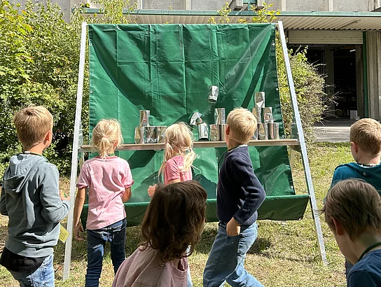 Mehrere Kinder stehen im Freien vor einer Spielstation mit aufgestapelten silberfarbenen Dosen. Einige Dosen befinden sich gerade im Fallen.