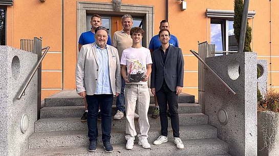 Sechs Männer stehen für ein Gruppenfoto auf der Treppe vor einem orangefarbenen Gebäude. Im Mittelpunkt steht der neue Auszubildende Leonhard Zenger, der einen Geschenkkorb in den Händen hält. Neben und hinter ihm stehen fünf Männer, die ihn zum Beginn seiner Ausbildung willkommen heißen.