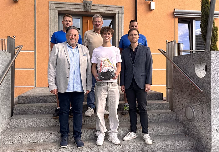 Sechs Männer stehen für ein Gruppenfoto auf der Treppe vor einem orangefarbenen Gebäude. Im Mittelpunkt steht der neue Auszubildende Leonhard Zenger, der einen Geschenkkorb in den Händen hält. Neben und hinter ihm stehen fünf Männer, die ihn zum Beginn seiner Ausbildung willkommen heißen.
