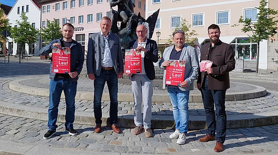 Fünf Personen stehen bei sonnigem Wetter auf einem Stadtplatz vor einem Brunnen beziehungsweise Denkmal und präsentieren rote Veranstaltungsplakate zum „16. Städtedreieckslauf“. Im Hintergrund sind Gebäude der Innenstadt zu sehen. Zwei der Personen halten zusätzlich Flyer in der Hand. Die Szene vermittelt den offiziellen Auftakt beziehungsweise die Vorstellung der Veranstaltung.