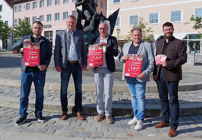 Fünf Personen stehen bei sonnigem Wetter auf einem Stadtplatz vor einem Brunnen beziehungsweise Denkmal und präsentieren rote Veranstaltungsplakate zum „16. Städtedreieckslauf“. Im Hintergrund sind Gebäude der Innenstadt zu sehen. Zwei der Personen halten zusätzlich Flyer in der Hand. Die Szene vermittelt den offiziellen Auftakt beziehungsweise die Vorstellung der Veranstaltung.