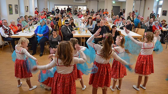 Mehrere Kinder tanzen in roten Kostümen mit blauen Tüchern vor einem großen, gut besuchten Saal voller sitzender und applaudierender Seniorinnen und Senioren.