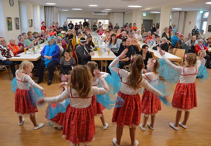 Mehrere Kinder tanzen in roten Kostümen mit blauen Tüchern vor einem großen, gut besuchten Saal voller sitzender und applaudierender Seniorinnen und Senioren.
