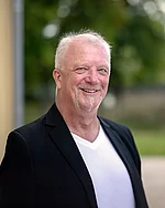 Ein älterer Mann mit sehr kurzen Haaren und markantem Lächeln trägt ein dunkles Sakko.
