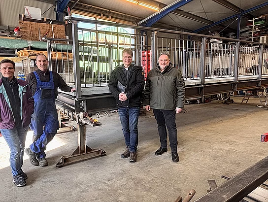 Vier Personen stehen in einer Werkhalle neben einer großen Metallbrücke, die dort aufgebaut und vermutlich für den Einbau vorbereitet wird.