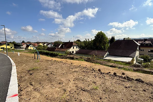 Ein leicht ansteigendes, unbebautes Grundstück mit brauner Erde liegt an einer neu asphaltierten Straße; im Hintergrund stehen einzelne Häuser am Waldrand unter blauem Himmel.