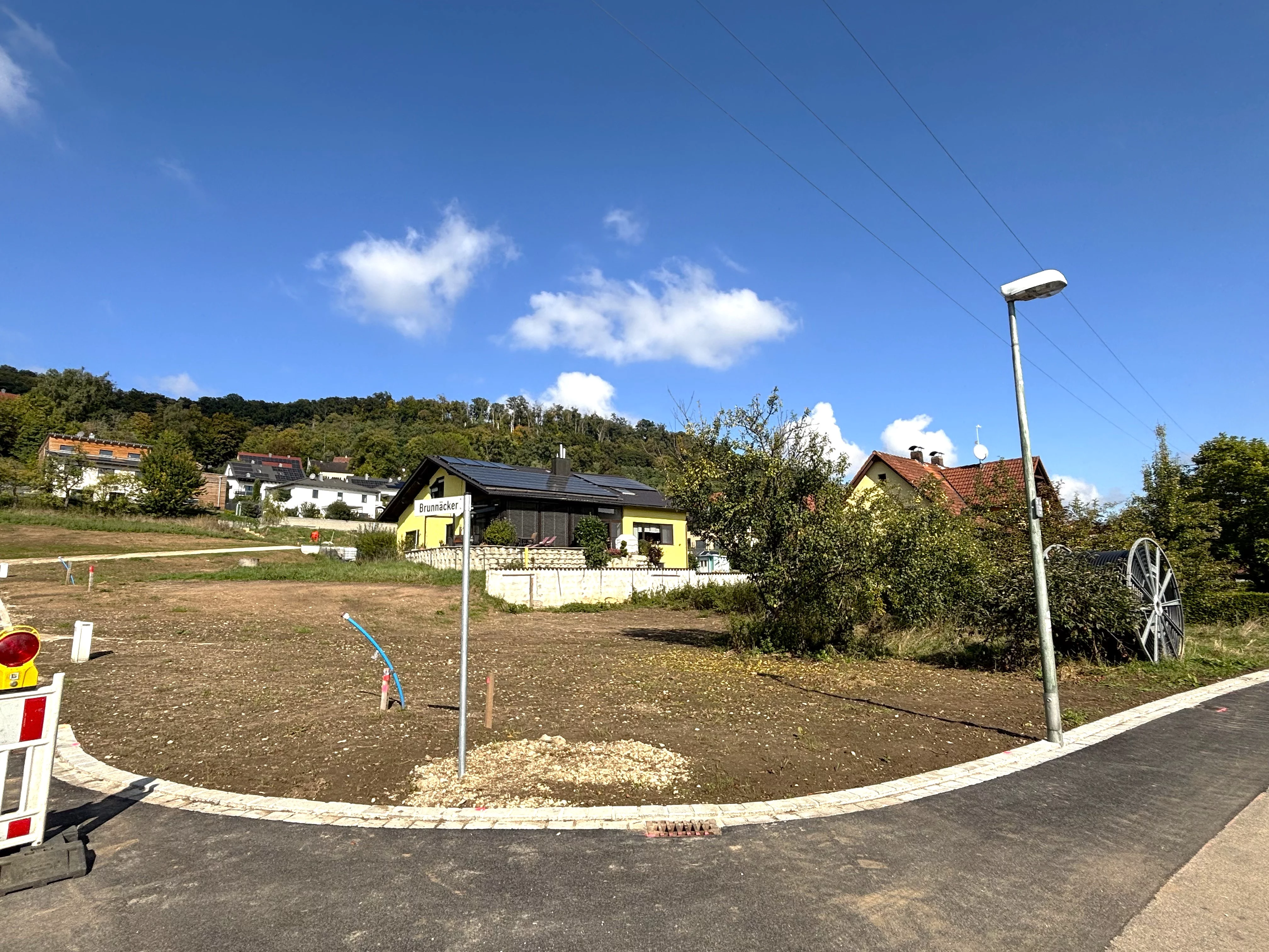 Ein leicht ansteigendes, unbebautes Grundstück mit brauner Erde liegt an einer neu asphaltierten Straße; im Hintergrund stehen einzelne Häuser am Waldrand unter blauem Himmel.
