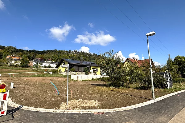 Ein leicht ansteigendes, unbebautes Grundstück mit brauner Erde liegt an einer neu asphaltierten Straße; im Hintergrund stehen einzelne Häuser am Waldrand unter blauem Himmel.