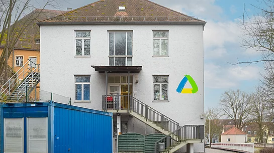 Vor einem zweigeschossigen weißen Gebäude mit zentralem Eingang und Außentreppe steht links ein großer blauer Container, während rechts an der Fassade ein buntes dreieckiges Logo angebracht ist.