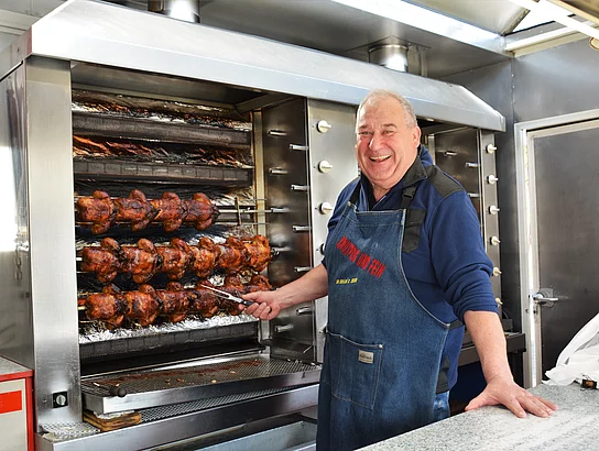 Ein Mann mit Grillzange in der Hand der vor einem Backhendl-Grill steht.