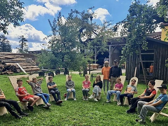 Eine Gruppe von Kindern sitzt im Freien im Halbkreis auf selbst gebauten Holzstühlen. Zwei Erwachsene stehen hinter der Gruppe; im Hintergrund sind Bäume, Holzstapel und Werkstattgebäude zu sehen.