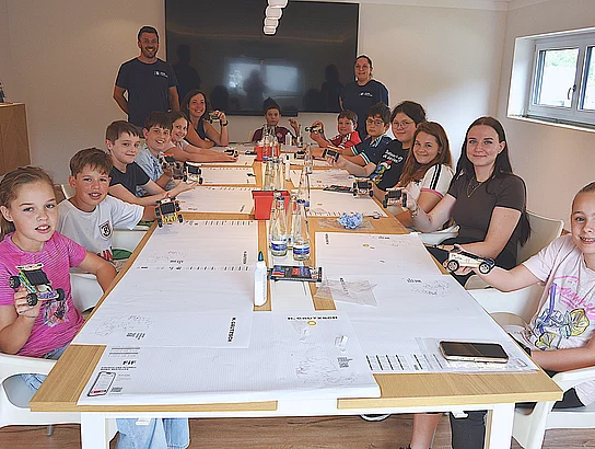 Kinder und Jugendliche sitzen gemeinsam mit Erwachsenen an einem langen Tisch. Mehrere von ihnen halten kleine selbst gebaute Fahrzeuge in die Kamera; auf dem Tisch liegen Arbeitsunterlagen und Bastelmaterialien.