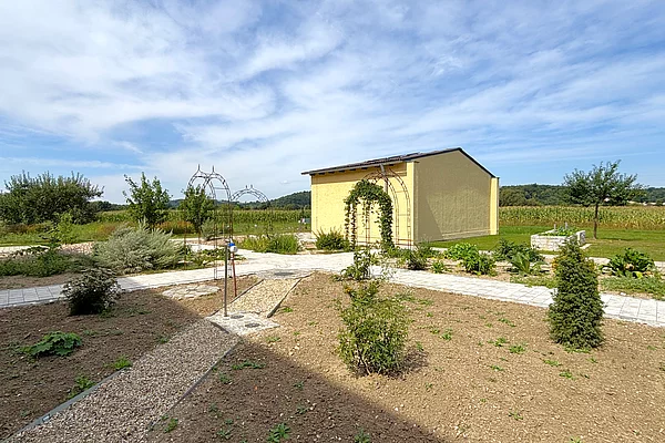 Ein mit Kieswegen angelegtes Naturbeet. Im Hintergrund das gelb gestrichene Gerätehaus des GOV Saltendorf.