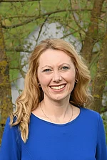 Eine Frau mit langen blonden Haaren und blauem Oberteil lächelt freundlich.