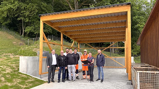Eine Gruppe von acht Personen steht unter einem neu errichteten, offenen Carport aus Holz mit Metall­dach auf einem gepflasterten Platz. Einige tragen Alltagskleidung, zwei Personen auffällige orangefarbene Arbeitskleidung. Im Hintergrund befinden sich ein bewaldeter Hang sowie technische Anlagen. Rechts sind große Metallcontainer zu sehen. Die Gruppe blickt in die Kamera und steht mittig unter der Konstruktion, die den Eindruck einer offiziellen Besichtigung oder Fertigstellung am Wasserwerk vermittelt.