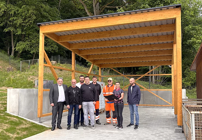 Eine Gruppe von acht Personen steht unter einem neu errichteten, offenen Carport aus Holz mit Metall­dach auf einem gepflasterten Platz. Einige tragen Alltagskleidung, zwei Personen auffällige orangefarbene Arbeitskleidung. Im Hintergrund befinden sich ein bewaldeter Hang sowie technische Anlagen. Rechts sind große Metallcontainer zu sehen. Die Gruppe blickt in die Kamera und steht mittig unter der Konstruktion, die den Eindruck einer offiziellen Besichtigung oder Fertigstellung am Wasserwerk vermittelt.